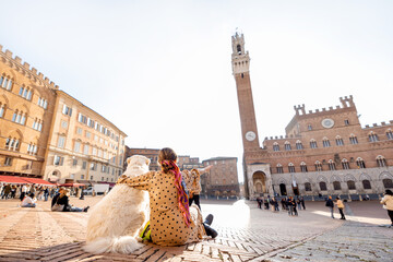 Obraz premium Woman sitting with her dog on main square of Siena city with a town hall on background. View from the backside. Concept of travel on tuscany and friendship with pet