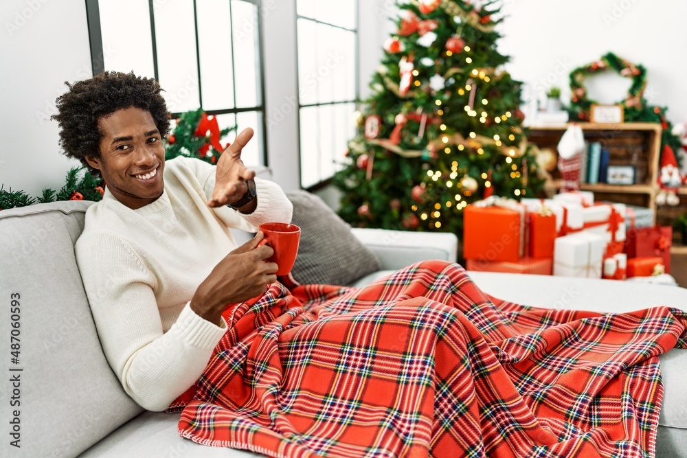 Sticker Young african american man sitting on the sofa drinking coffee by christmas tree smiling cheerful offering palm hand giving assistance and acceptance.