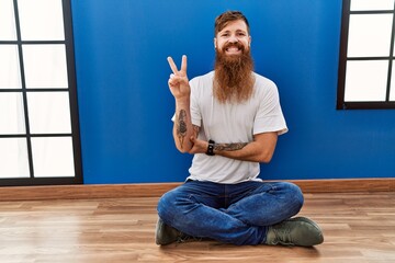 Redhead man with long beard sitting on the floor at empty room smiling with happy face winking at...