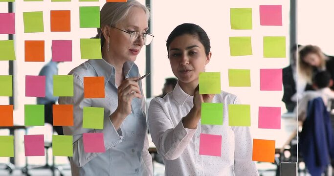Mature lady mentor young Indian female intern create workflow model by sticking colored memo cards on glass office board. Two diverse women coworkers develop business process using agile kanban method