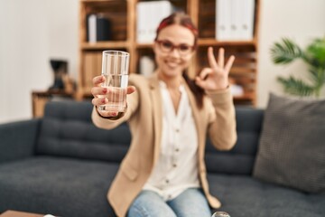 Young caucasian woman offering a glass of water doing ok sign with fingers, smiling friendly gesturing excellent symbol