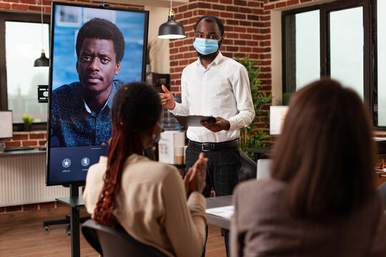 Multi-ethnic teamwork with medical face masks against covid19 discussing financial statistics with company leader during online videocall meeting conference in startup office. Business call on monitor