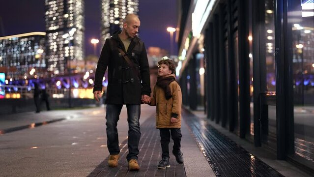 father and son are walking along a blurry evening street against the background of city lights. they hold hands and chat. the camera is moving. the general plan