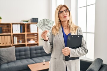 Middle age blonde woman working at therapy consultation office holding money smiling looking to the side and staring away thinking.