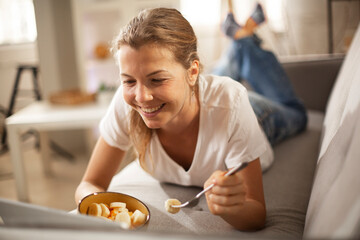 Cheerful young woman eating healthy breakfast. Beautiful woman lying on sofa, eating fruit