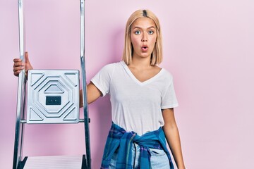 Beautiful blonde woman holding ladder scared and amazed with open mouth for surprise, disbelief face