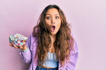 Young hispanic girl holding bowl of sugar candy scared and amazed with open mouth for surprise,...