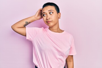 Beautiful hispanic woman with short hair wearing casual pink t shirt confuse and wondering about question. uncertain with doubt, thinking with hand on head. pensive concept.