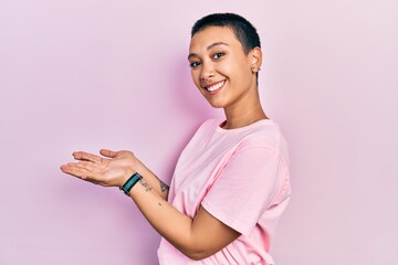 Beautiful hispanic woman with short hair wearing casual pink t shirt pointing aside with hands open palms showing copy space, presenting advertisement smiling excited happy