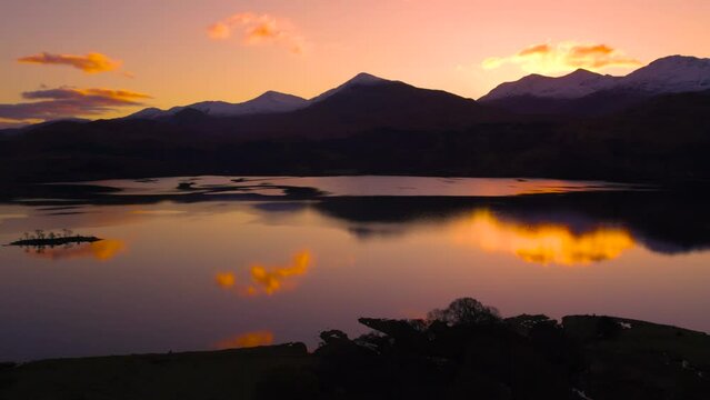 Aerial drone footage slowly rising to reveal Glen Etive and Loch Etive in Scotland during a fiery orange sunrise, with snow-capped mountains, silhouetted trees and still water reflecting clouds.