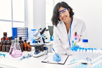 Fototapeta premium Young latin woman wearing scientist uniform holding test tube writing on clipboard at laboratory