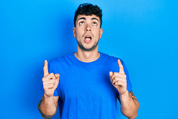 Fototapeta premium Young hispanic man wearing casual blue t shirt amazed and surprised looking up and pointing with fingers and raised arms.