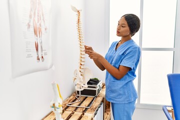 Young latin woman wearing physiotherapist uniform pointing with finger to vertebral column at clinic