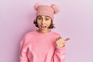 Young brunette woman wearing cute wool cap smiling with happy face looking and pointing to the side with thumb up.