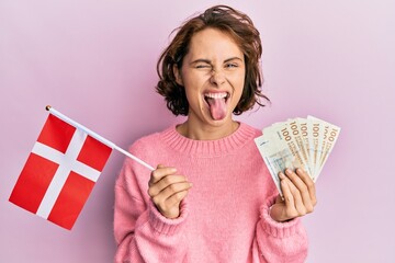 Young brunette woman holding norway flag and krone banknotes sticking tongue out happy with funny...