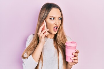 Young blonde girl using smartphone and drinking a cup of coffee angry and mad screaming frustrated and furious, shouting with anger. rage and aggressive concept.