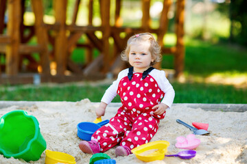 Cute toddler girl playing in sand on outdoor playground. Beautiful baby in red gum trousers having fun on sunny warm summer day. Child with colorful sand toys. Healthy active baby outdoors plays games