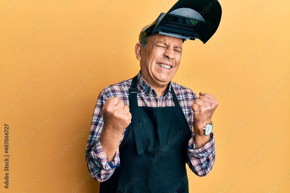 Poster Handsome senior man with grey hair wearing welding protection mask very happy and excited doing winner gesture with arms raised, smiling and screaming for success. celebration concept.