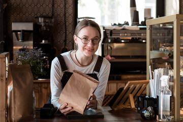 Fototapeta premium Young smiling woman (salesperson in bakery) holding craft paper packing bag for taking away food in coffee shop
