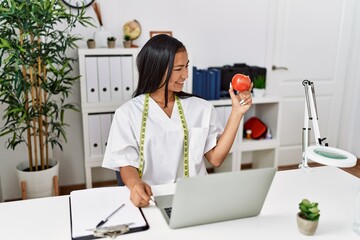 Young latin woman wearing nutritionist uniform holding apple at clinic