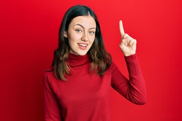 Young brunette woman wearing turtleneck sweater smiling with an idea or question pointing finger up with happy face, number one