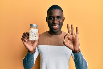 Young african american man holding jar with pumpkin seeds doing ok sign with fingers, smiling friendly gesturing excellent symbol