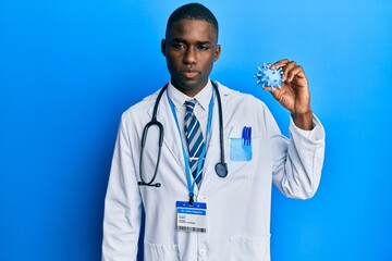 Young african american man wearing doctor coat holding virus toy thinking attitude and sober expression looking self confident