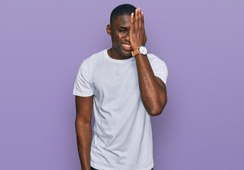 Young african american man wearing casual white t shirt covering one eye with hand, confident smile on face and surprise emotion.