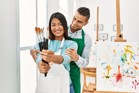 Young latin painter couple smiling happy holding paintbrushes at art studio