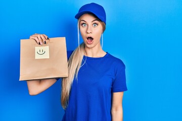 Young caucasian woman holding take away paper bag with happy face reminder scared and amazed with open mouth for surprise, disbelief face