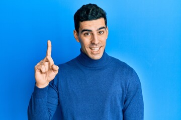 Naklejka premium Handsome hispanic man wearing turtleneck sweater smiling with an idea or question pointing finger up with happy face, number one