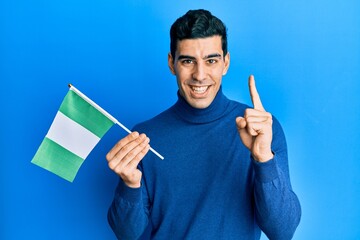 Handsome hispanic man holding nigeria flag smiling with an idea or question pointing finger with happy face, number one