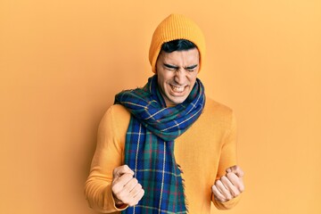 Handsome hispanic man wearing wool hat and winter scarf very happy and excited doing winner gesture...