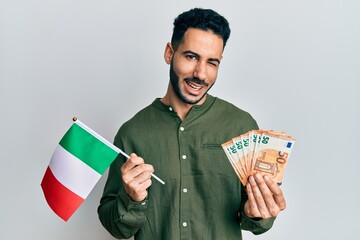 Young hispanic man holding italy flag and euros banknotes winking looking at the camera with sexy...
