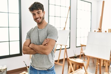 Young hispanic artist man smiling happy standing with arms crossed gesture at art studio.