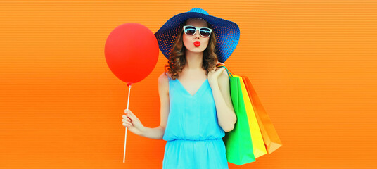 Portrait of beautiful happy young woman blowing her lips with shopping bags wearing summer straw hat on orange background
