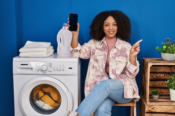Young african american woman doing laundry using smartphone smiling happy pointing with hand and finger to the side