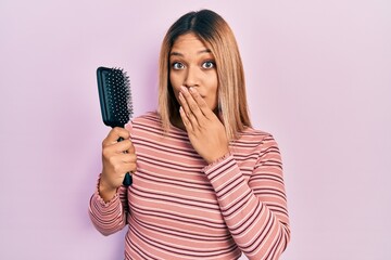 Beautiful hispanic woman holding hairbrush covering mouth with hand, shocked and afraid for...