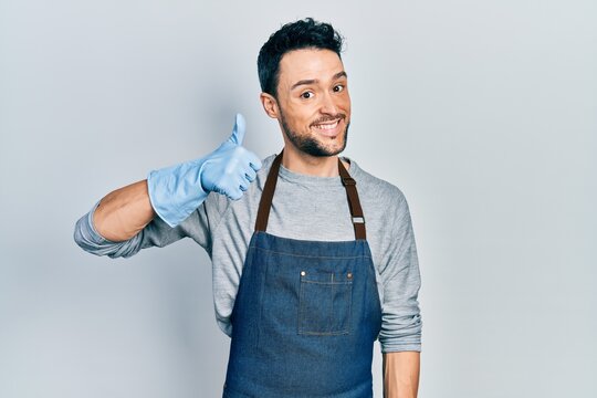 Young hispanic man wearing apron and gloves smiling happy and positive, thumb up doing excellent and approval sign