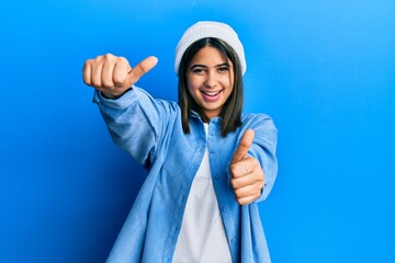 Young latin woman wearing cute wool cap approving doing positive gesture with hand, thumbs up smiling and happy for success. winner gesture.
