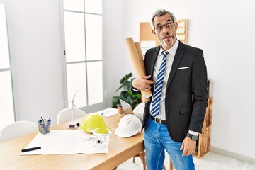 Middle age hispanic business man holding paper blueprints making fish face with lips, crazy and comical gesture. funny expression.