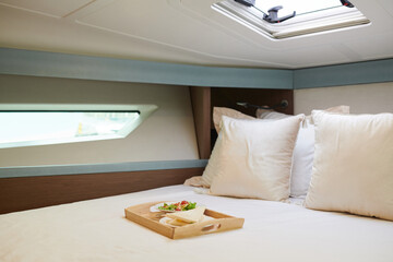 Tray with delicious vegetable salad and sandwich for lunch on bed in yacht cabin