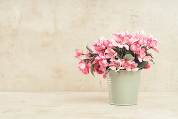 Pink spring flowers in a tiny olive bucket. Pastel beige cozy setup with copy space. Blooming fruit tree branch. Greeting card