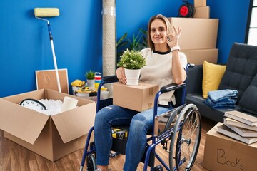 Young woman sitting on wheelchair moving to a new home smiling positive doing ok sign with hand and fingers. successful expression.