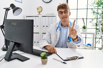 Young caucasian doctor man working at the clinic showing and pointing up with fingers number two while smiling confident and happy.