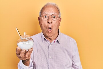Senior man with grey hair holding bowl with sugar scared and amazed with open mouth for surprise, disbelief face