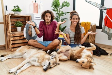 Young hispanic couple doing laundry with dogs clueless and confused expression with arms and hands raised. doubt concept.
