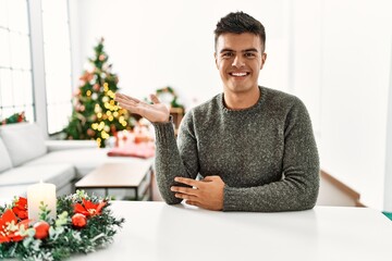 Young hispanic man sitting on the table by christmas tree smiling cheerful presenting and pointing with palm of hand looking at the camera.