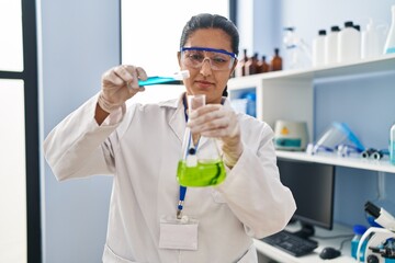 Young hispanic woman wearing scientist uniform pouring liquid on test tube at laboratory