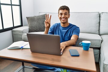 Young handsome hispanic man using laptop sitting on the floor showing and pointing up with fingers number three while smiling confident and happy.
